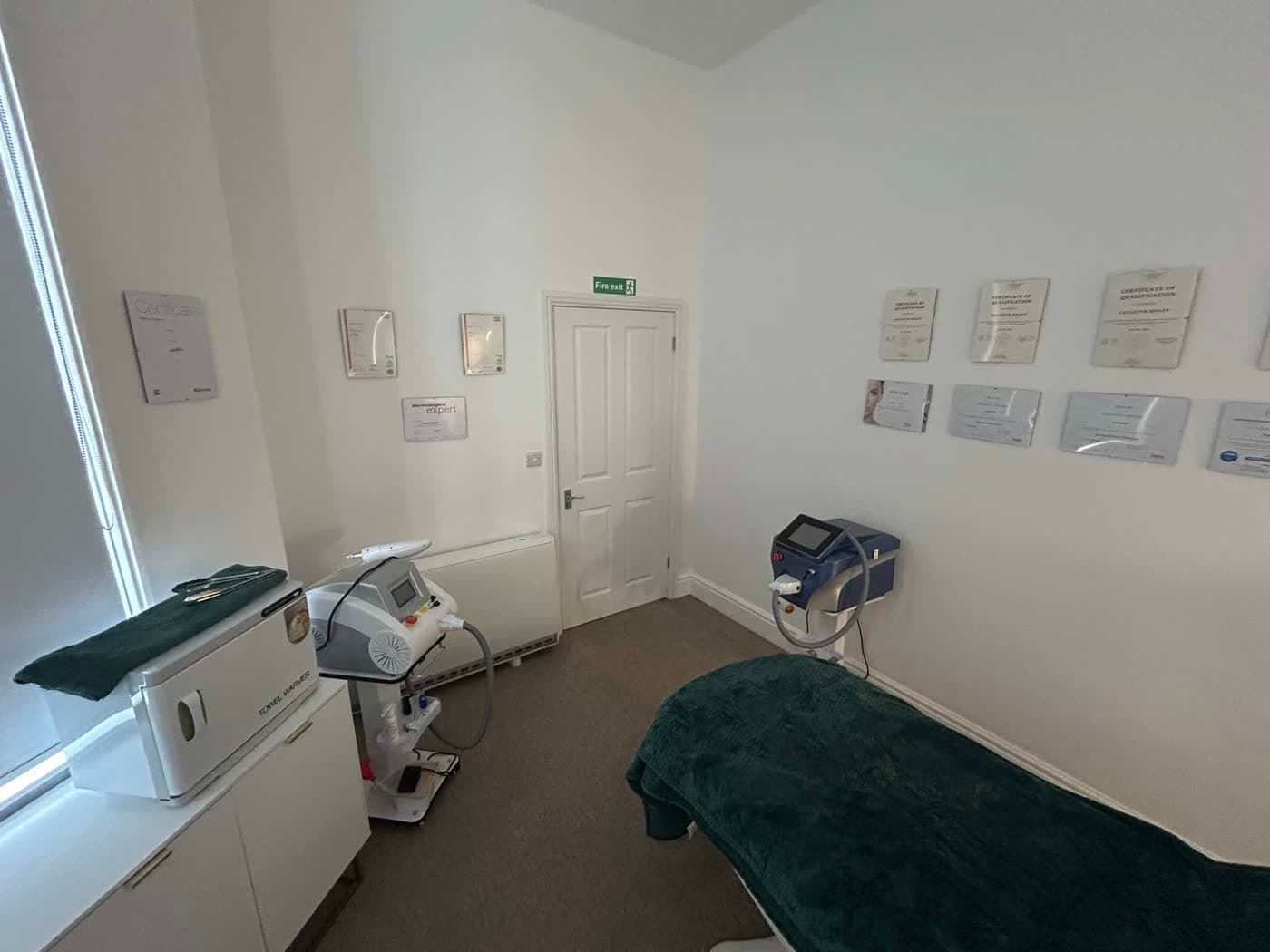 Treatment room with sage-throw bed, aesthetic-equipment trolley and Collette's certificates on display — including Dermalogica Expert.