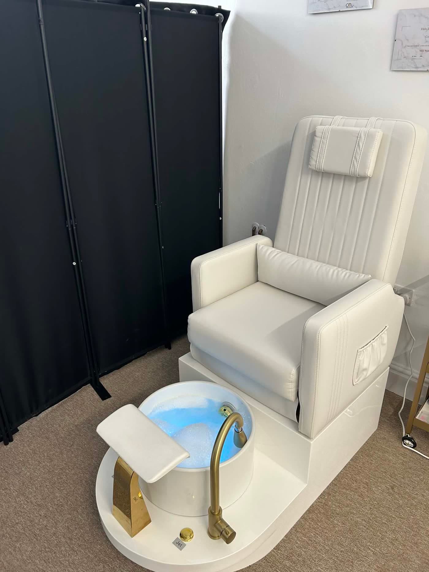 Pedicure spa station — white reclining chair with pillow and gold-tap foot bath, dark privacy screen behind.